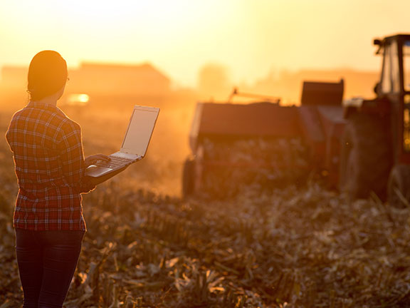 A woman standing in field using a laptop for tractor baler